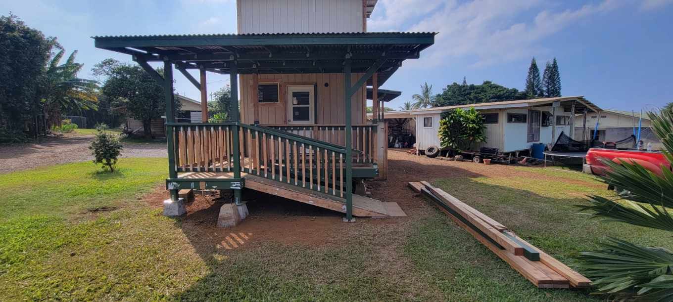 Small house with a green porch and awning. Boards in front of the house and another building in the background.