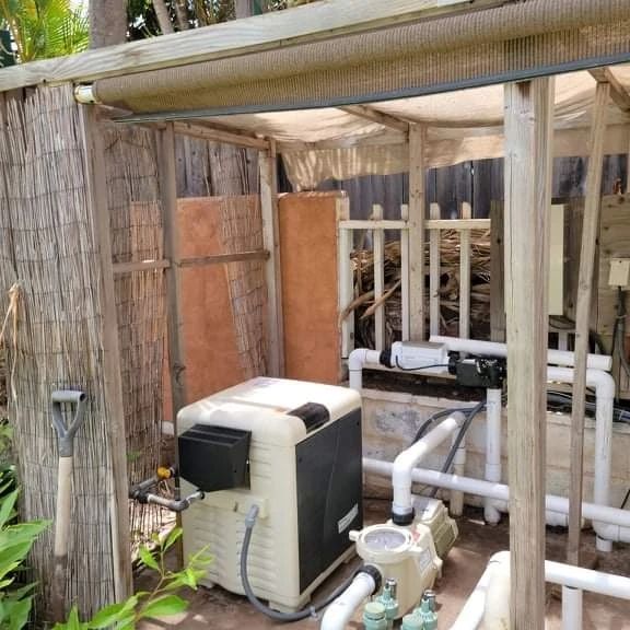 Pool equipment enclosure with a heater, pump, and plumbing. Wooden structure, beige and white colors.
