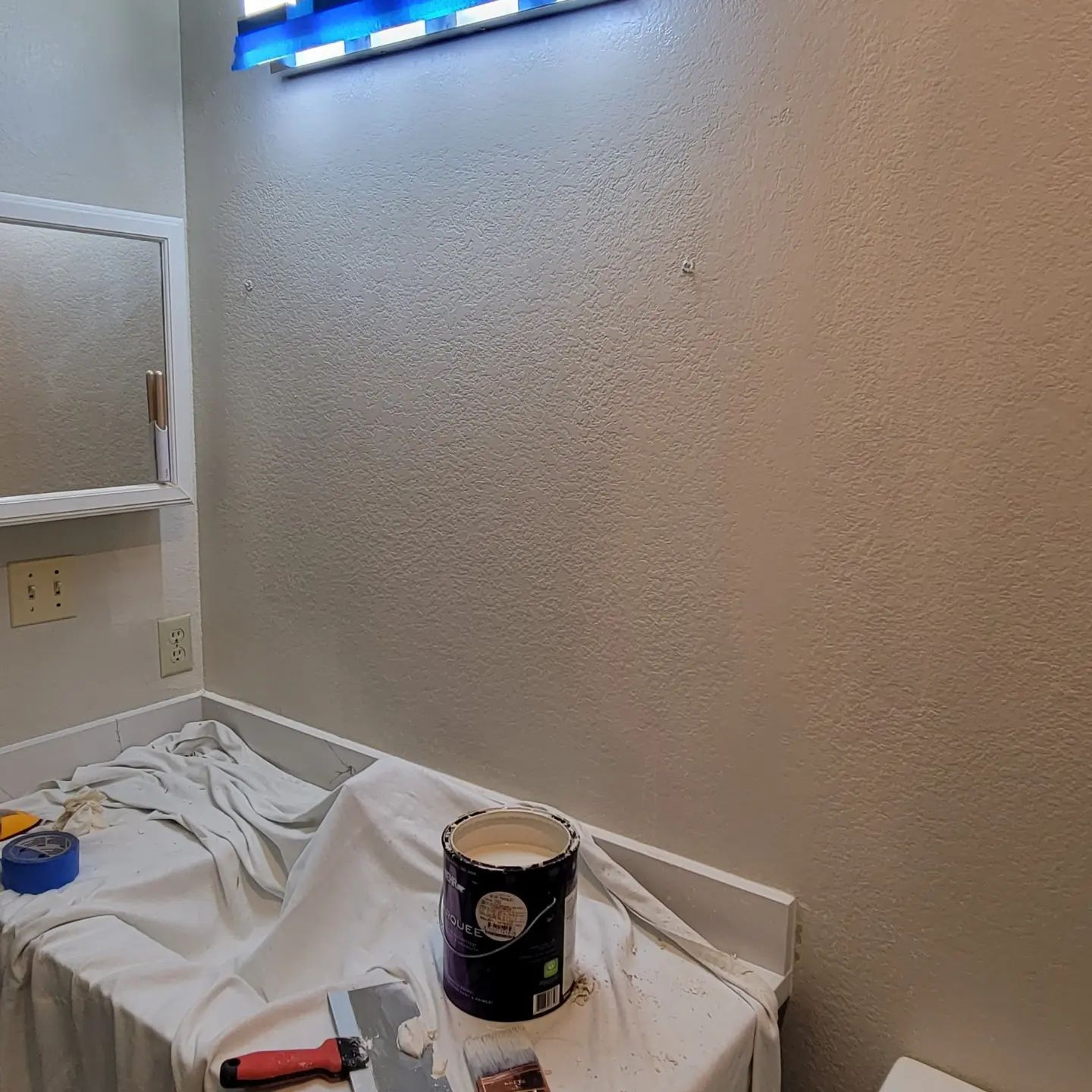 Bathroom with textured beige walls, a white countertop, mirror, and paint can.