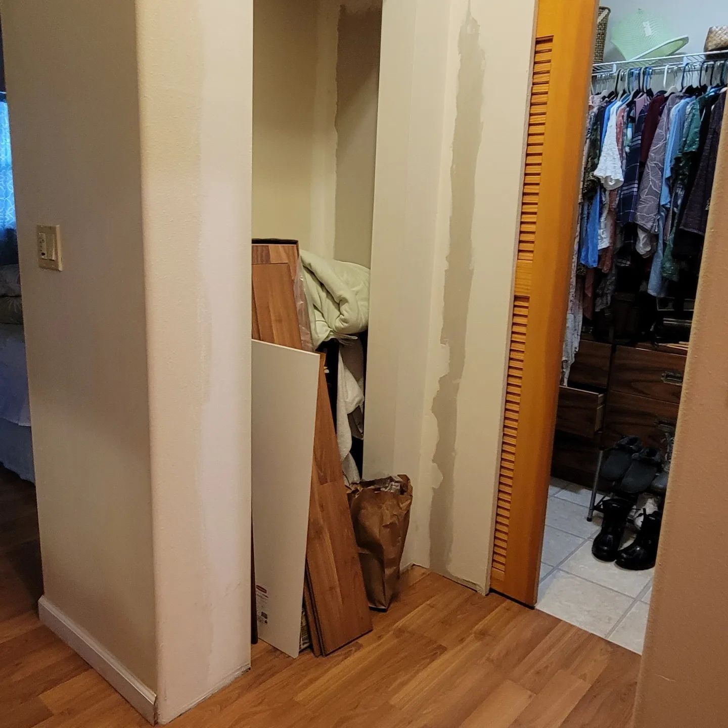 A small closet entrance with building materials inside. A wooden floor. Clothes hanging in the adjacent closet.