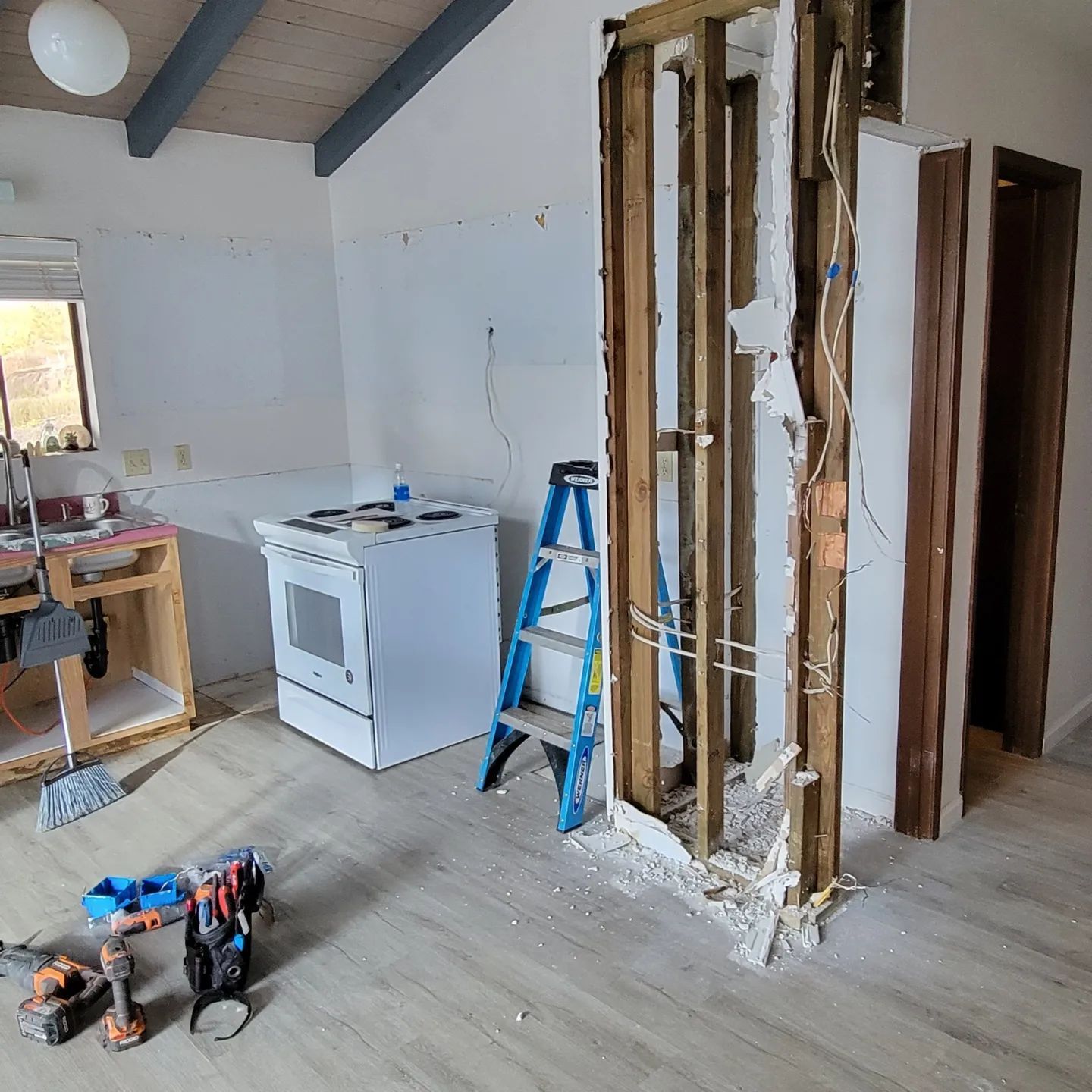 Kitchen remodel in progress: white stove, exposed wall studs, blue ladder, tools on floor.