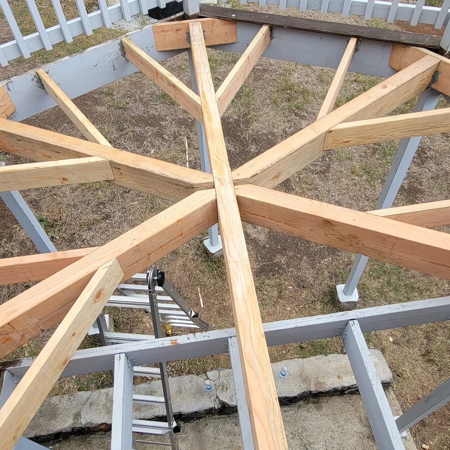 Overhead view of a deck frame with radial beams, some lumber, and a ladder on a concrete base.