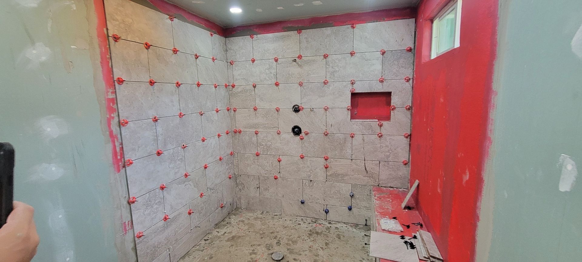 Interior view of a shower under construction. Red waterproofing membrane covers the walls; gray tiling material is installed.