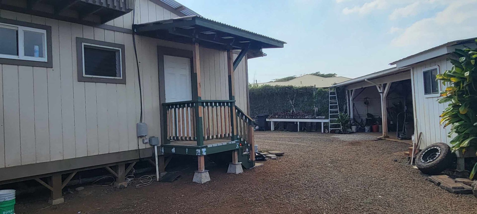 Exterior view of two single-story buildings with a gravel yard, a porch, and a lush green hedge.