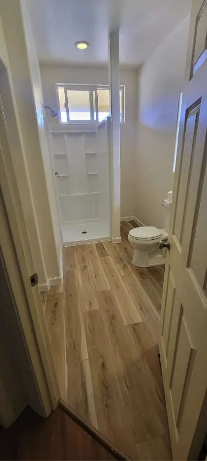 Narrow bathroom with wood floor, white shelving, toilet, and window at the far end.