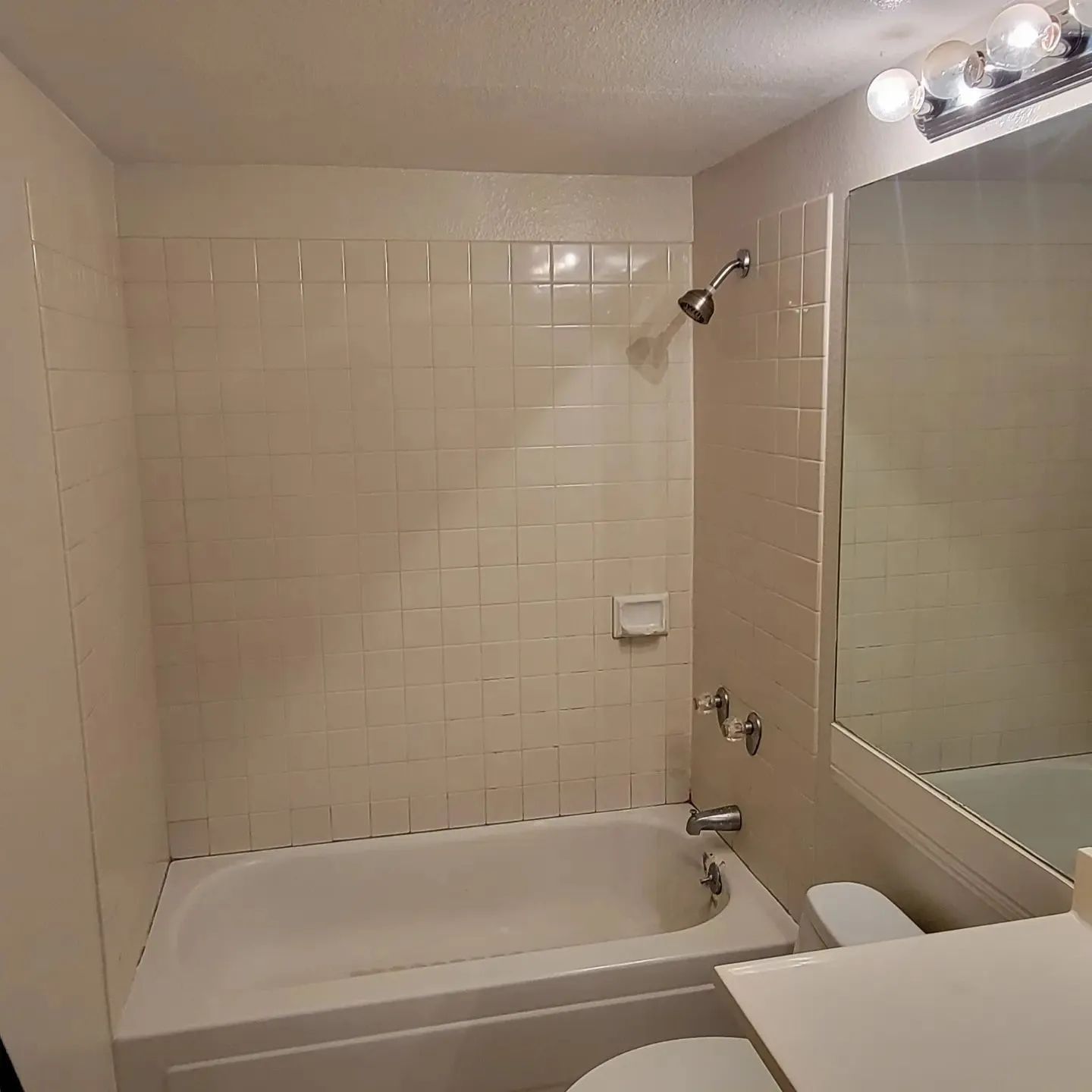 Small beige-tiled bathroom with a white bathtub, mirror, and sink counter under lights