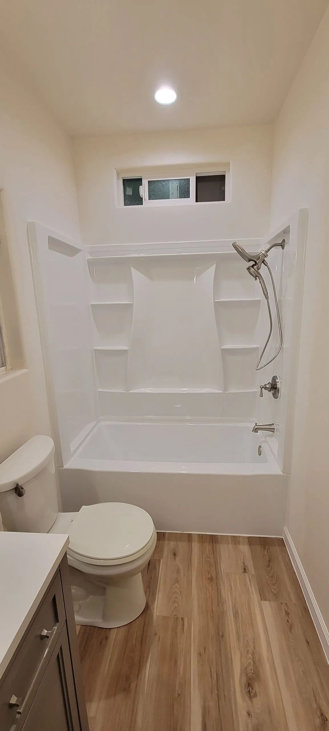 Small bathroom with wood-look floor, bathtub, toilet, sink, showerhead, shelves, and window above the tub
