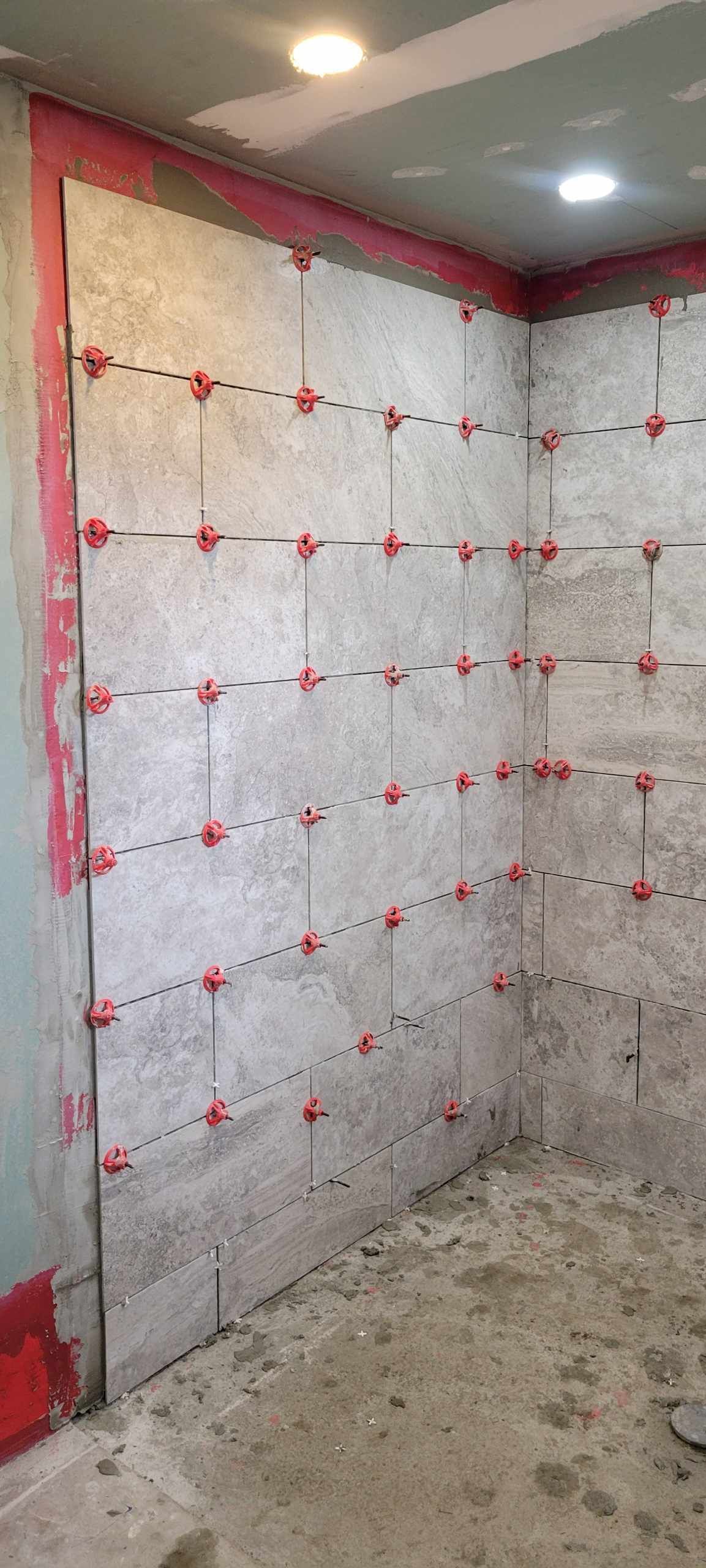 Tiles installed on interior wall, secured with red leveling clips, construction in progress.