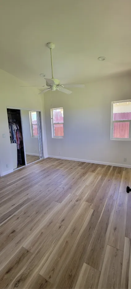 Empty room with light wood floor, white walls, ceiling fan, and windows with pink-tinted light.
