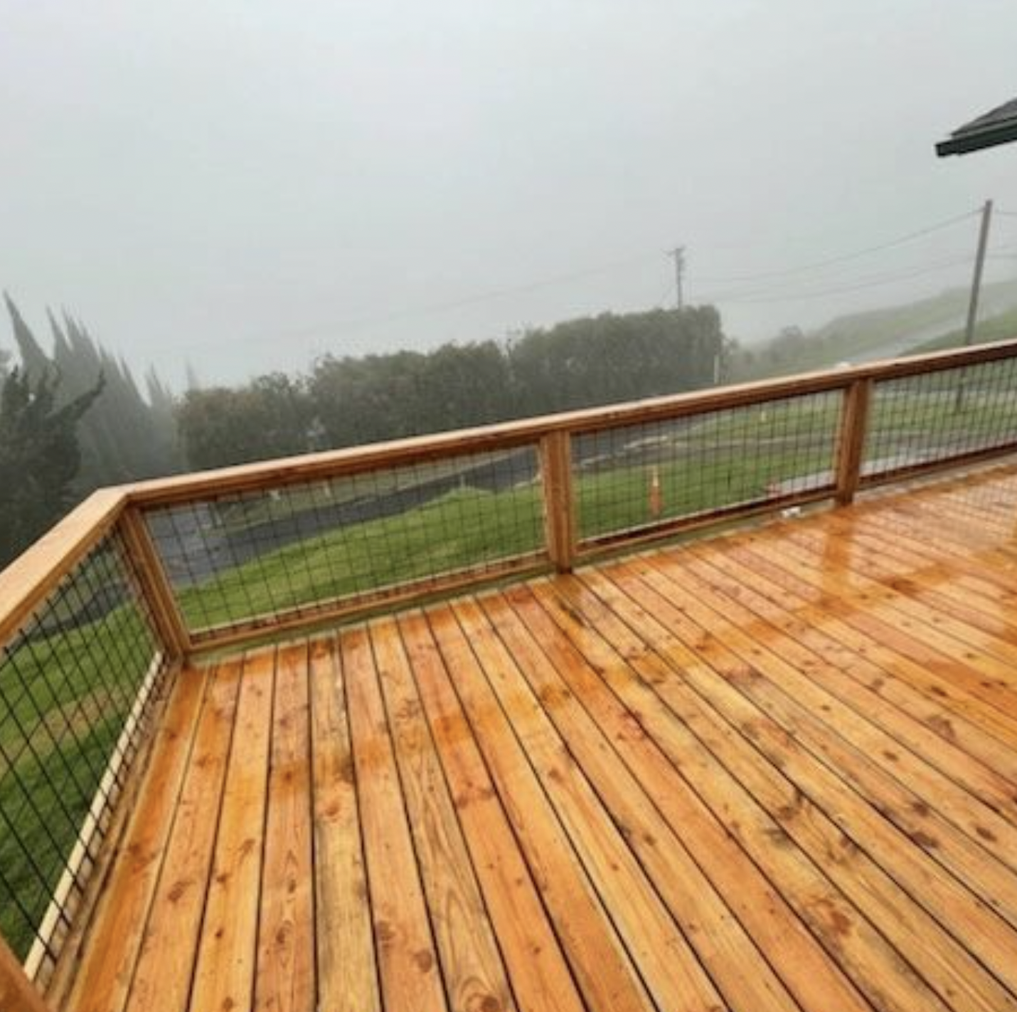 Wooden deck with wet, reddish-brown planks, wire railing, and a foggy green landscape in the background.
