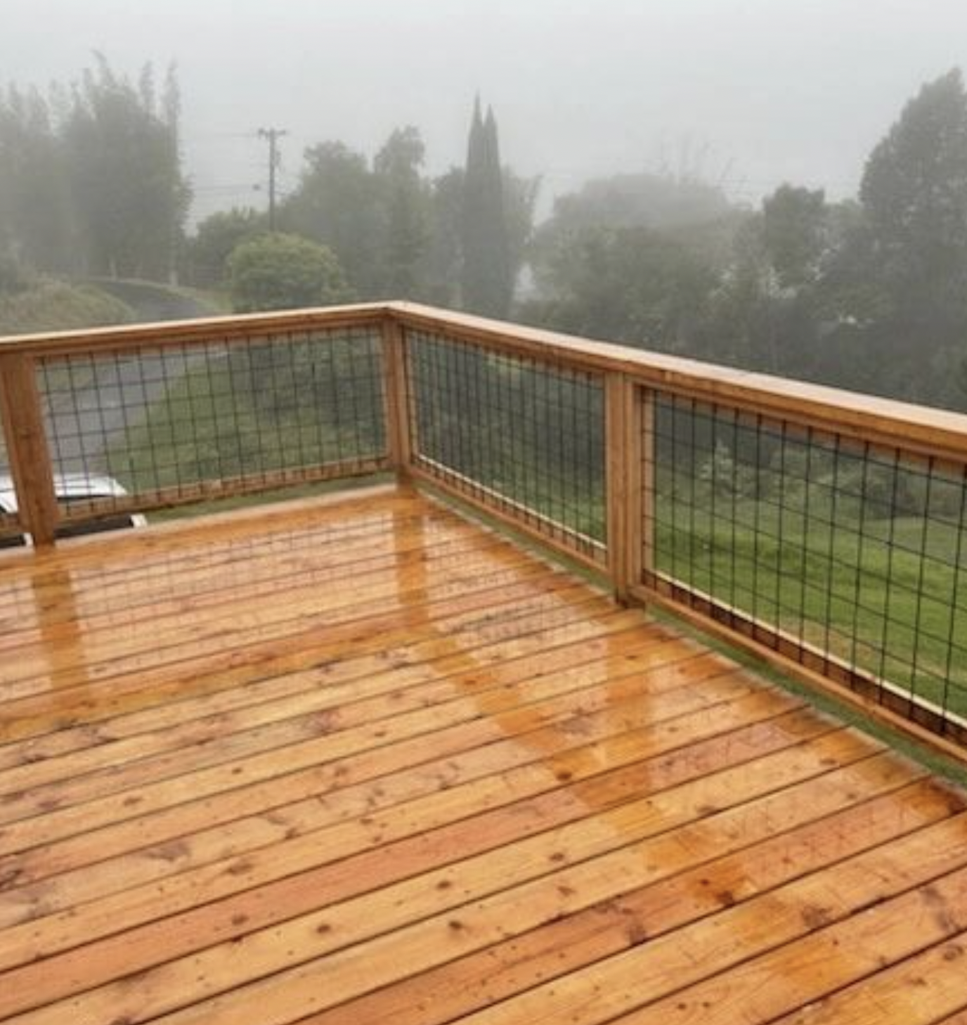 Wooden deck with wire mesh railing, wet from rain, overlooking a foggy landscape.