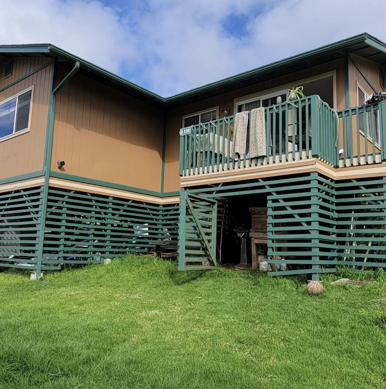 Brown house with green lattice base and deck, set on grassy lawn under a cloudy sky.