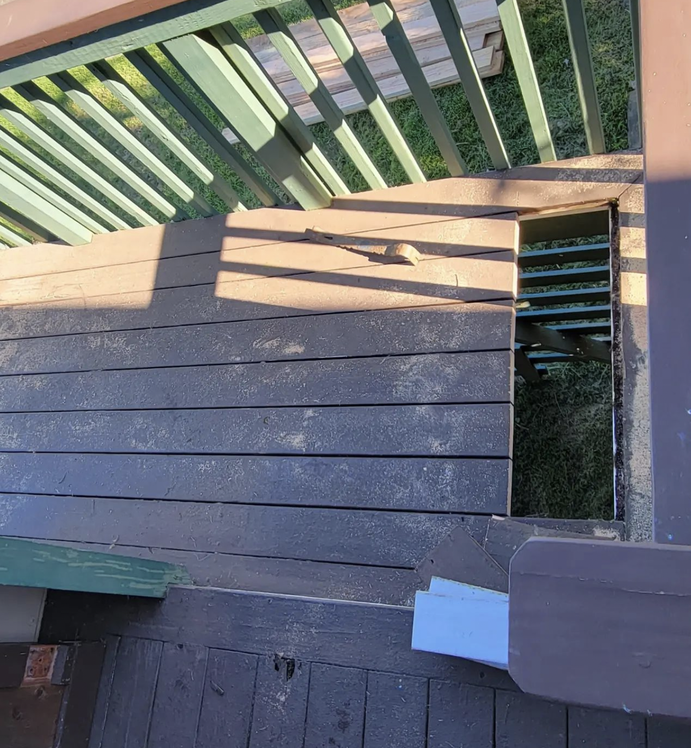 Wooden deck with a cut-out section and green railing above. Sunlight casts shadows.