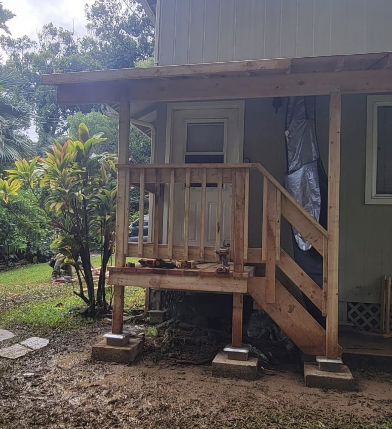Wooden porch with stairs leading to a door; concrete supports, railing, and small roof.