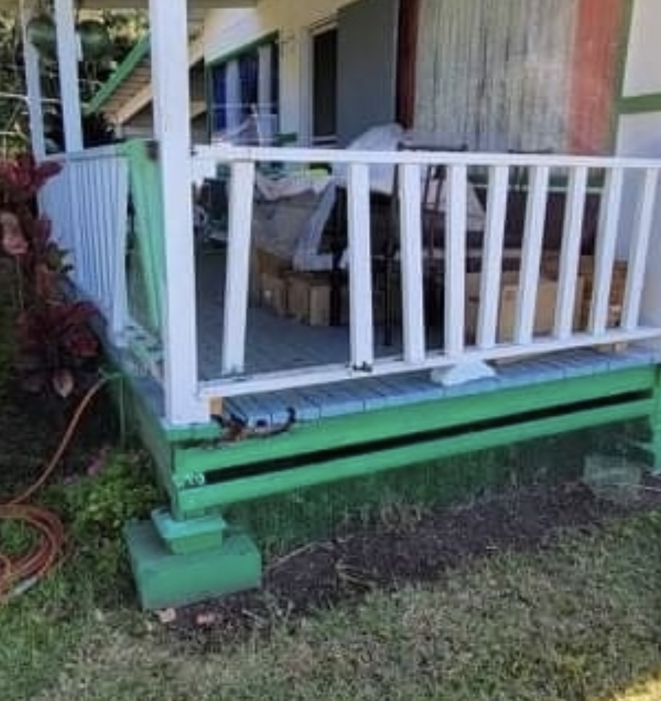 Green and white porch with railing. Some boxes on the porch, green painted support beams and a hose.