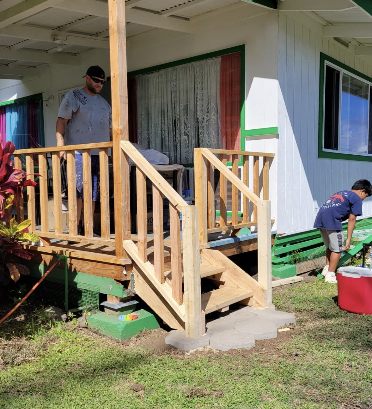 Man on a porch with newly built wooden stairs; child helping. House is white with green trim and colorful curtains.