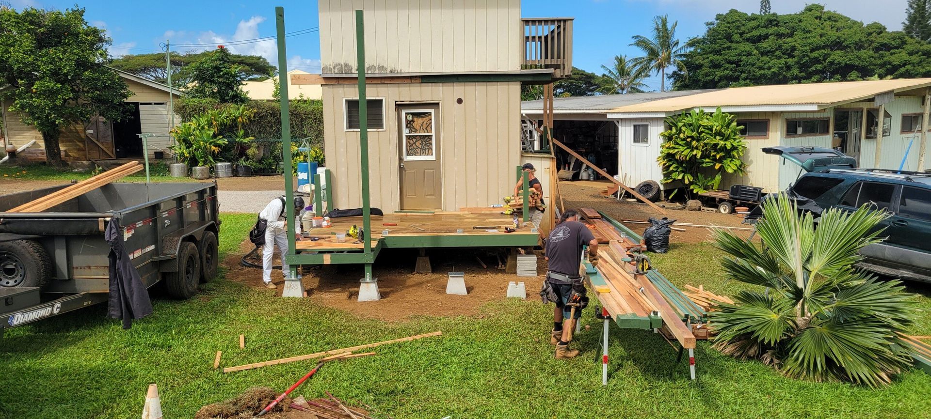 Construction workers building an outdoor deck next to a two-story building on a sunny day.
