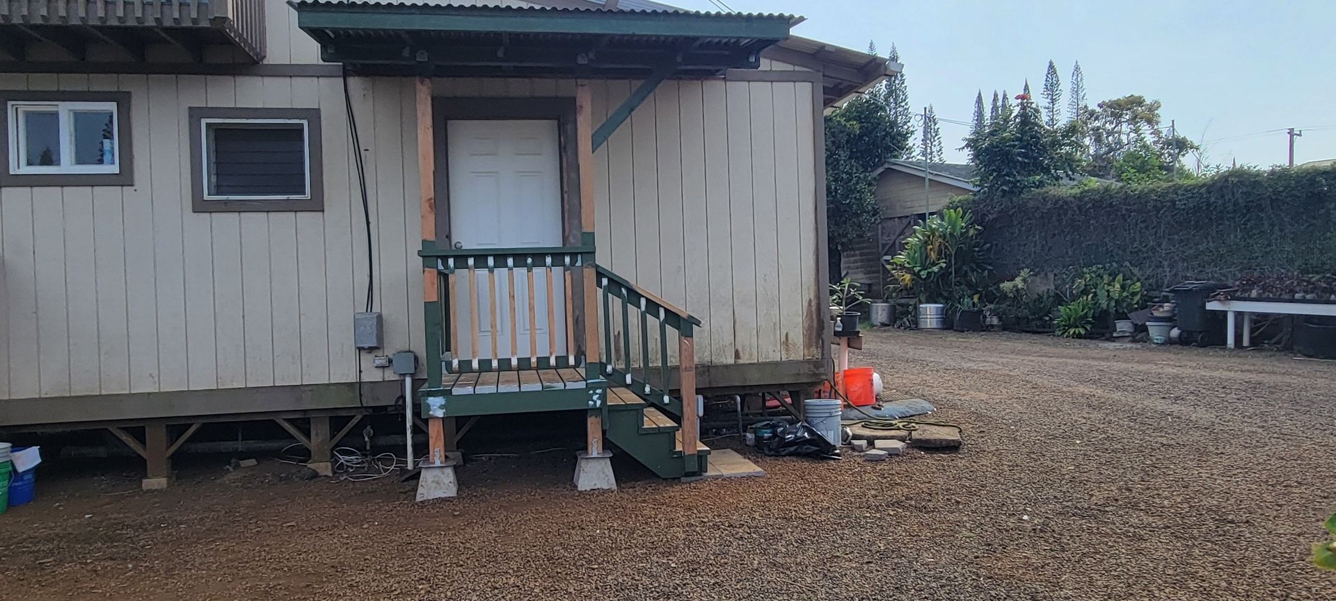 A weathered building with a porch, surrounded by gravel. A green hedge and potted plants are in the background.