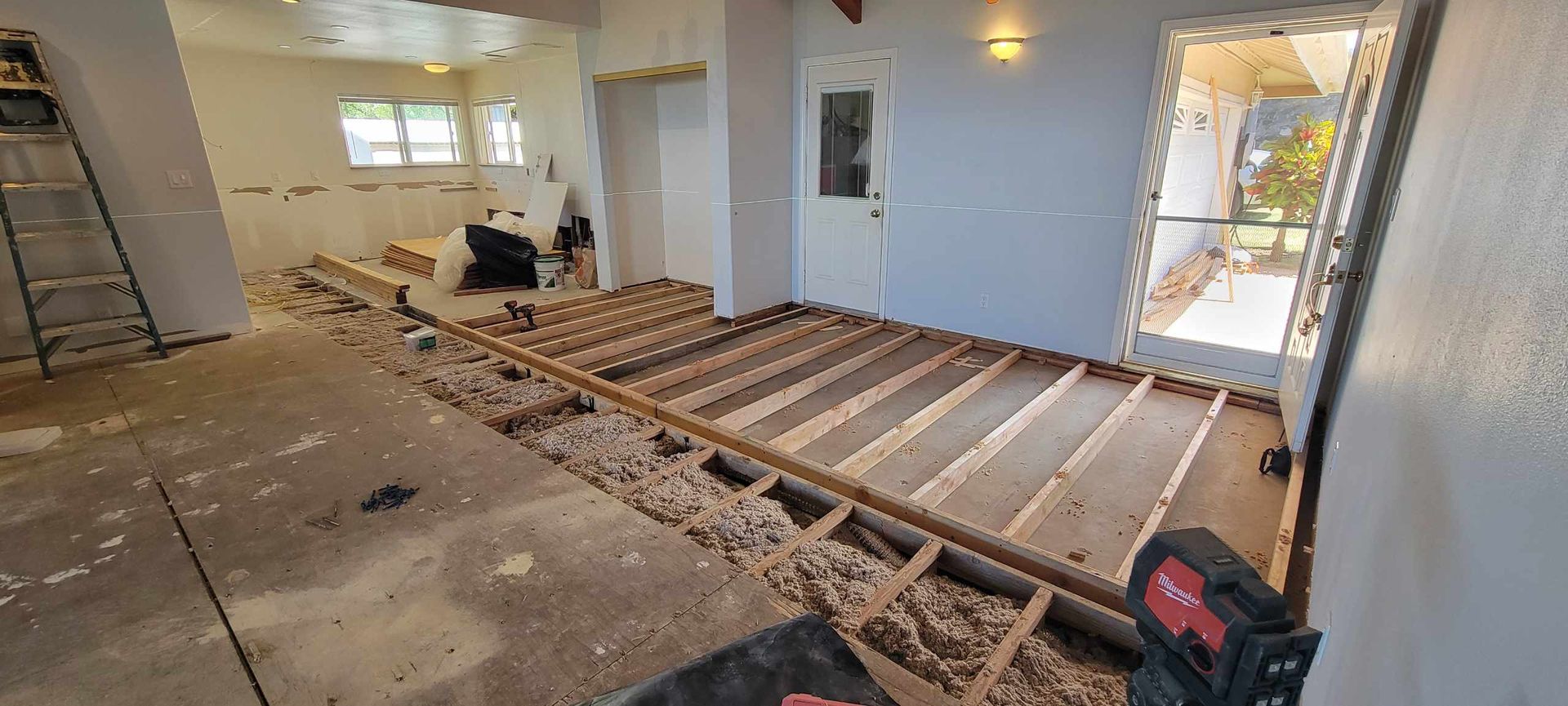 A room undergoing renovation; wooden floor frame exposed, open doorway to outside, ladder on left.