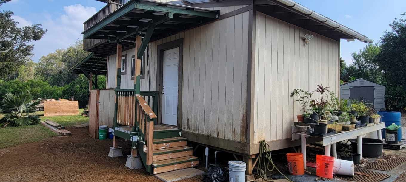 A small, weathered, single-story house with a porch and steps, surrounded by plants and outdoor items.