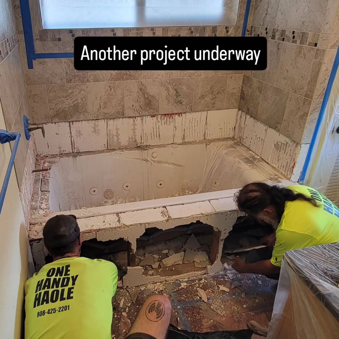 Workers demolish a tiled bathroom wall and tub during renovation, with debris scattered on the floor.