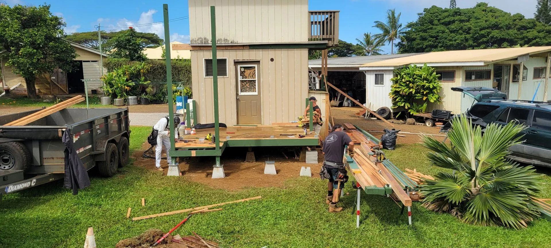 Workers assembling a small beige house module on a grassy site with trucks and tools nearby
