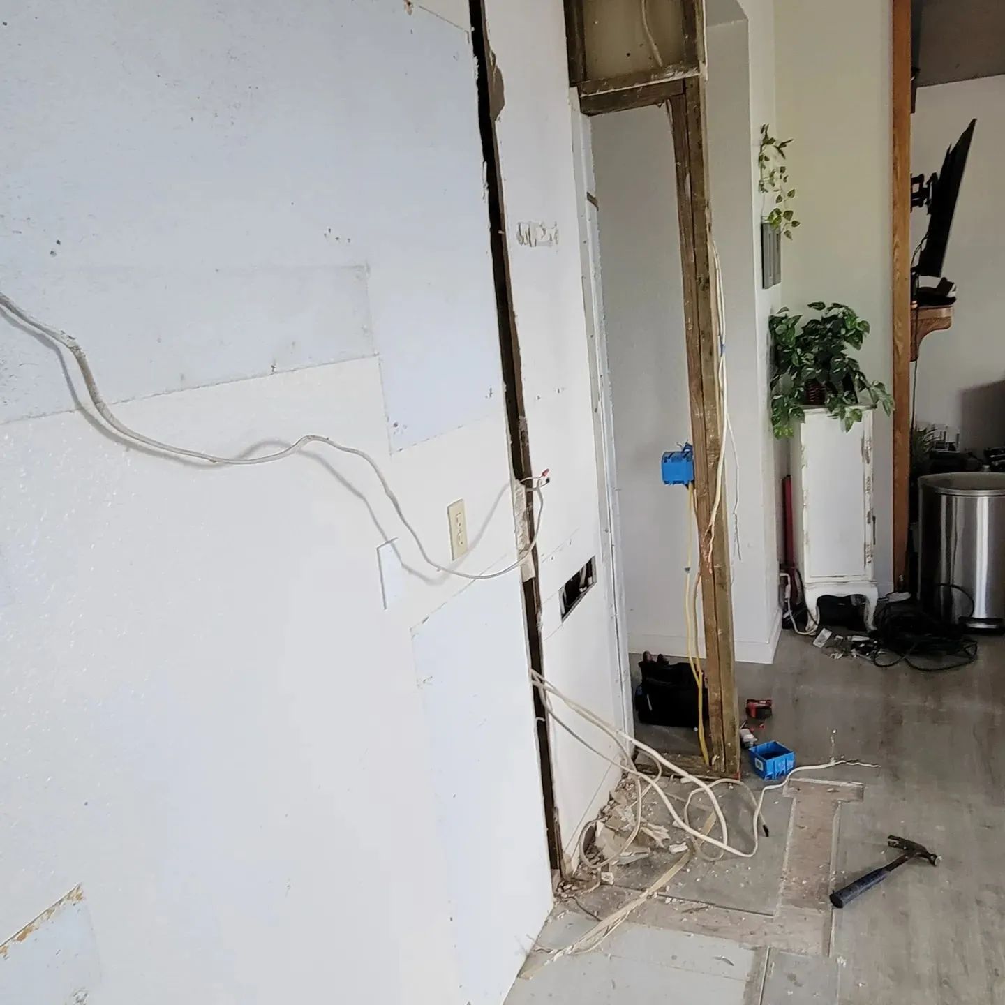Messy indoor hallway with exposed wires, a white wall, and tools on the floor near a doorway.