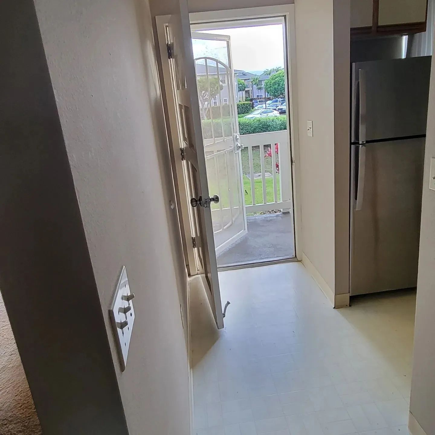Kitchen hallway with open front door, balcony view, and a refrigerator on the right.
