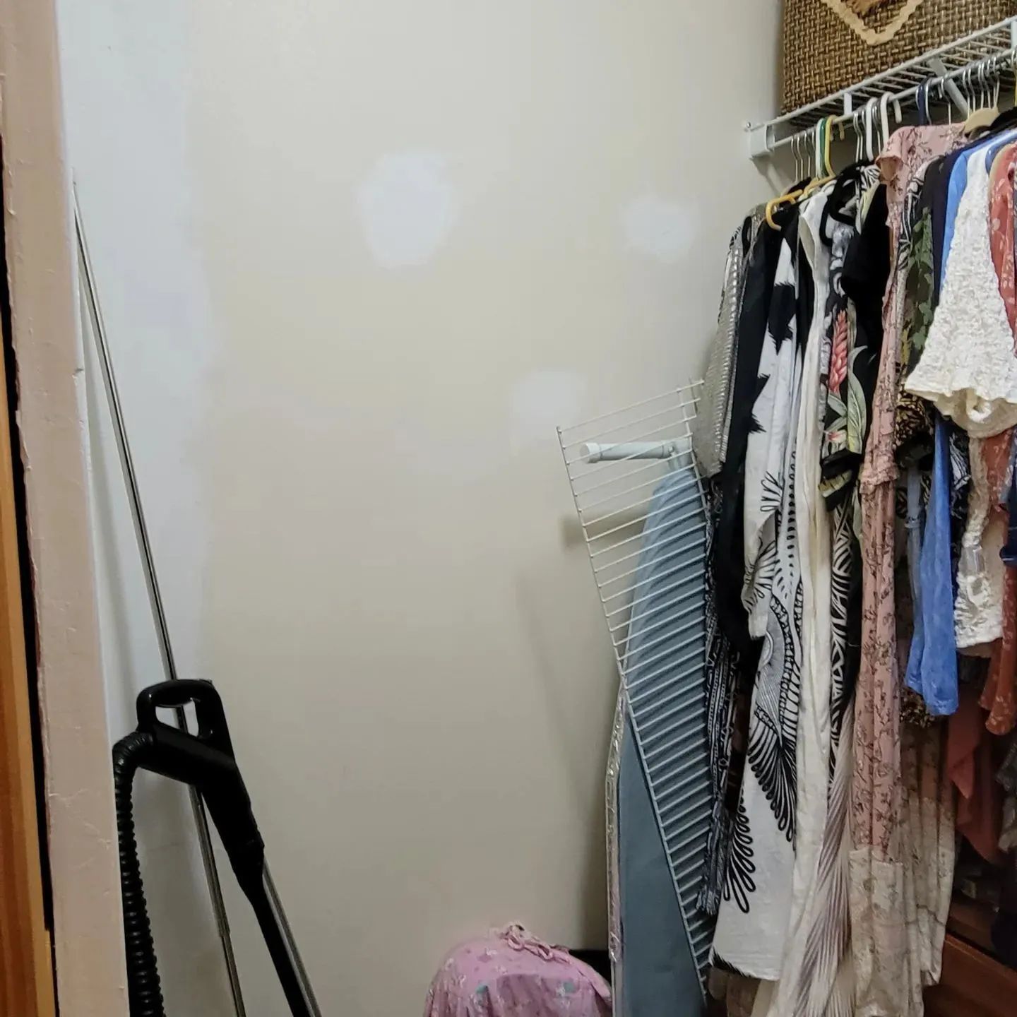 Small closet with hanging clothes, an ironing board, and a pink bag on the floor.