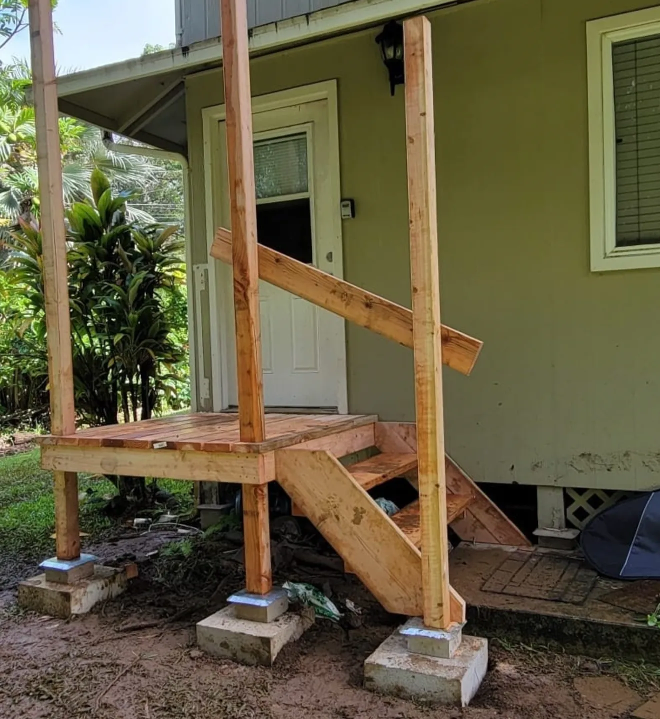 Wooden front porch stairs under construction beside a green house, with temporary posts and concrete footings