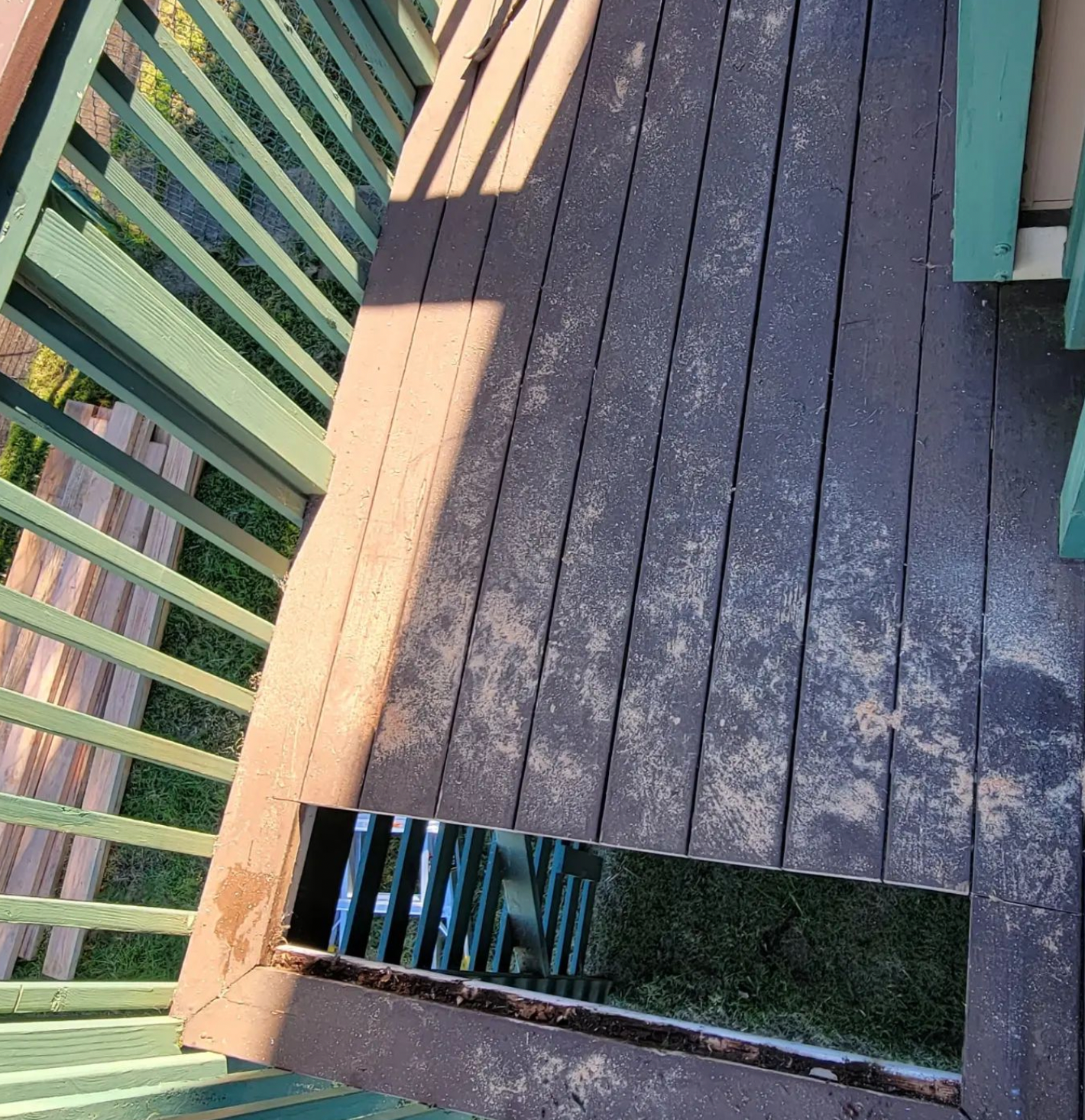 Wooden deck with green railing and a dark rectangular floor hatch, lit by sunlight