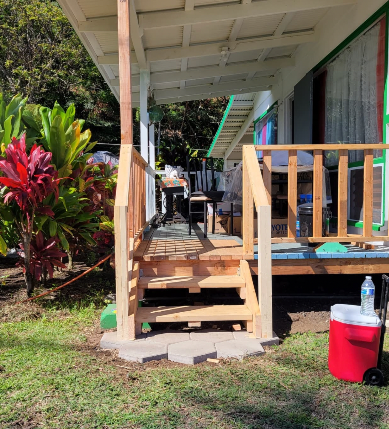 Small wooden porch with steps and railing beside a lawn, with a red cooler and water bottle in front.