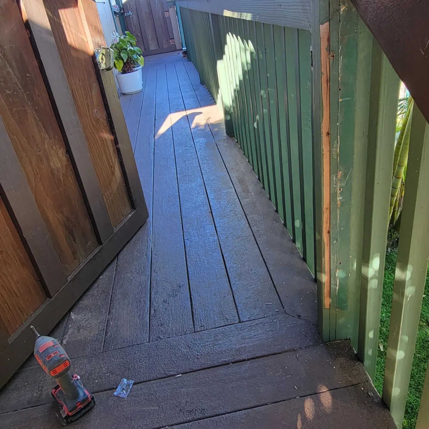 Narrow wooden balcony with green railing, potted plants, and a drill on the floor in sunlight.