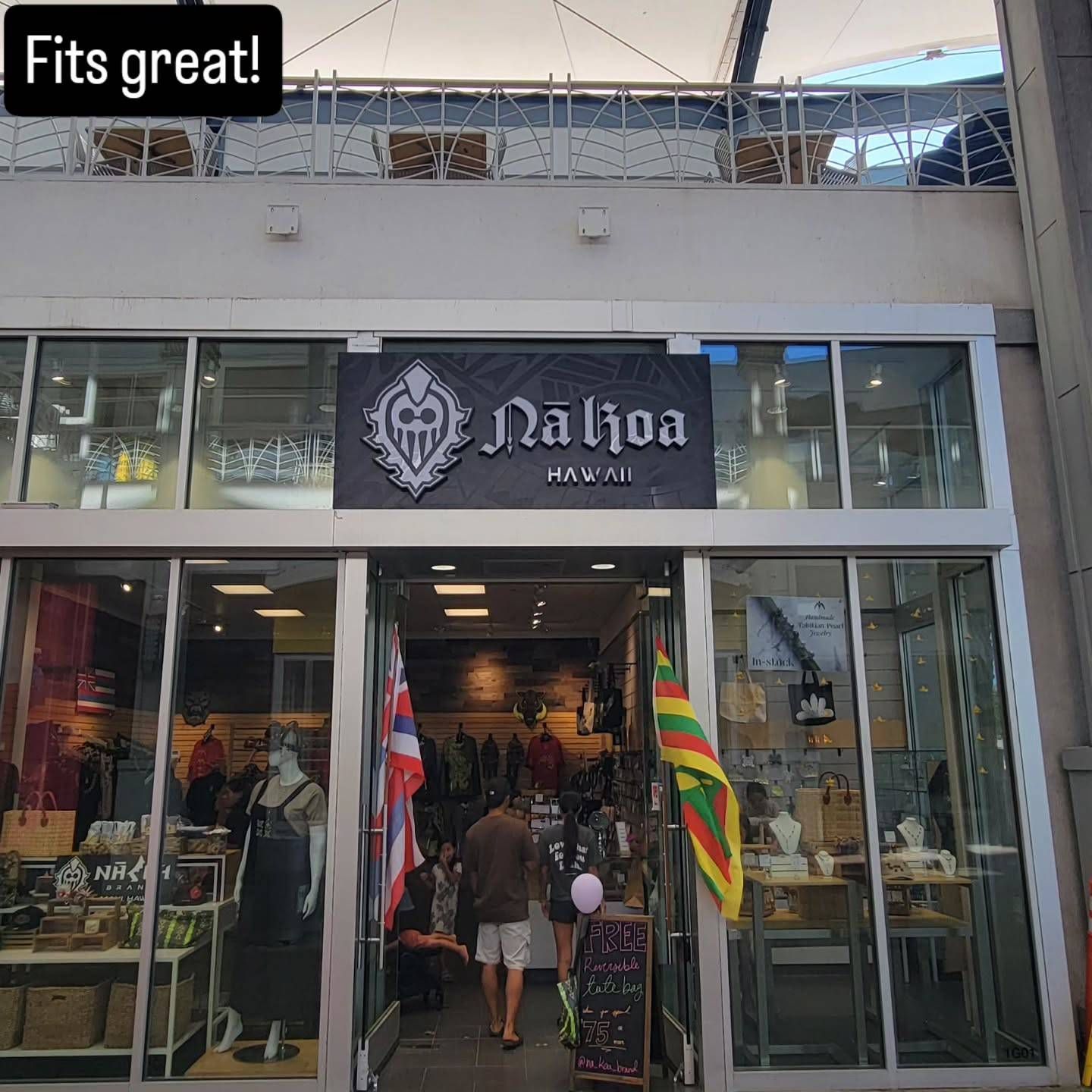 Exterior of Nā Likoa Hawai'i store. A person enters, Hawaiian flags flank the doorway, and merchandise is visible.