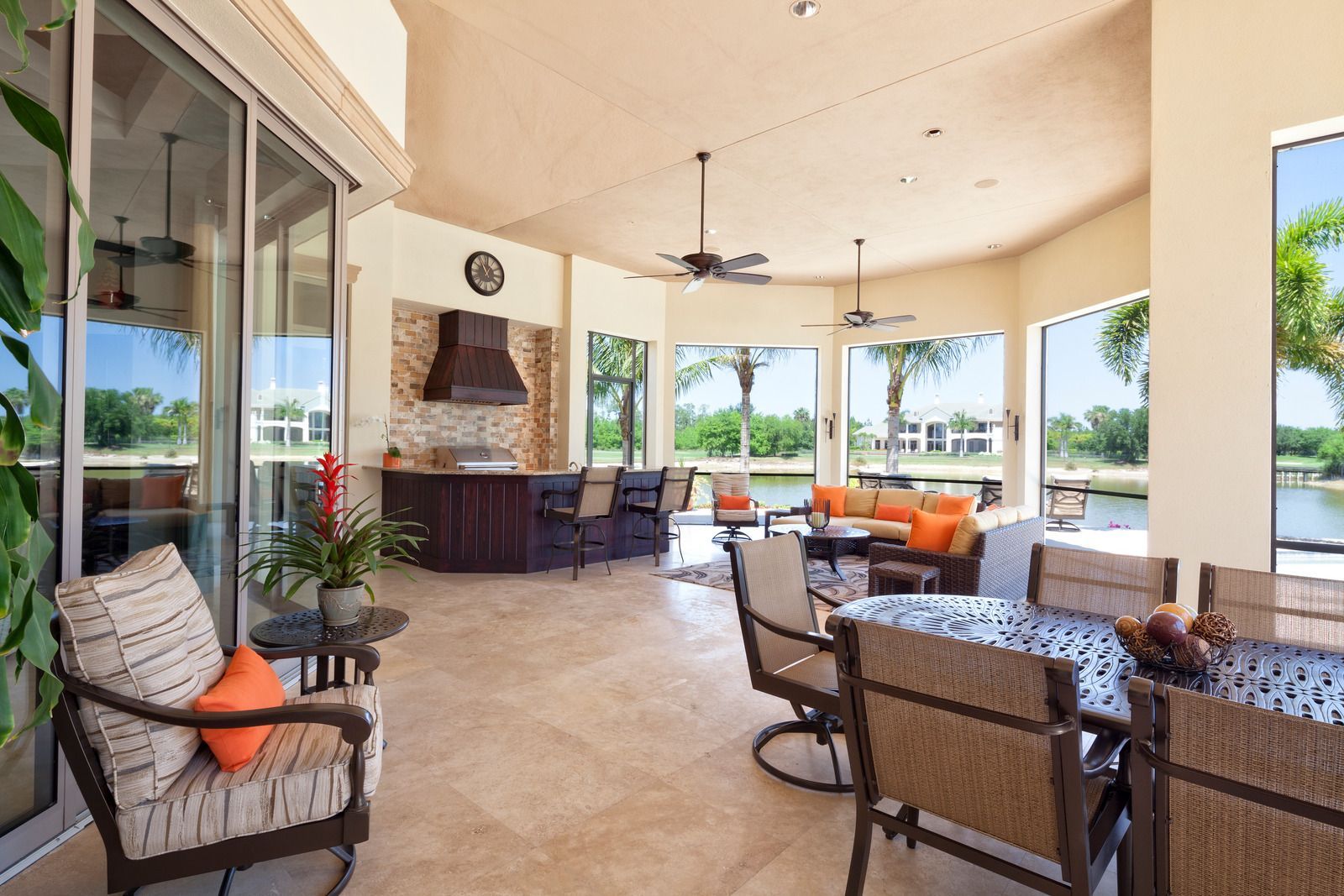 Spacious outdoor patio featuring a dining area, a lounge space with orange cushions, and a kitchen bar overlooking a lake.