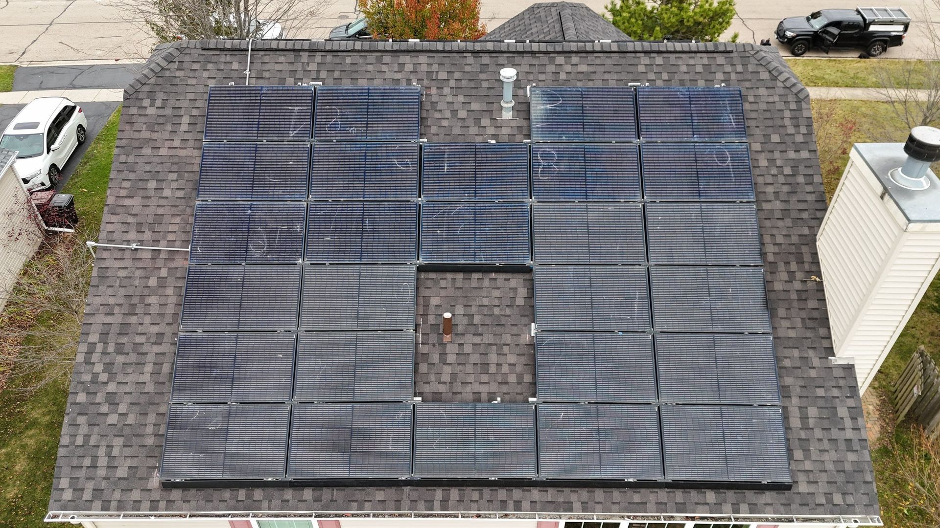 Solar panels installed on a dark shingle roof, covering most of the surface, with a chimney visible.