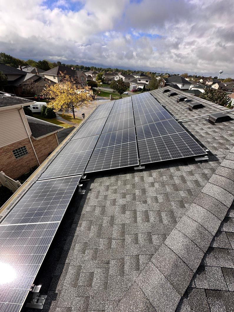 Solar panels on a shingled roof with neighborhood houses in the background under a cloudy sky.