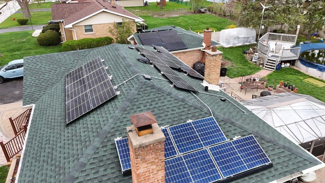 Green shingled roof with solar panels. Houses and yard in the background. Cloudy sky.