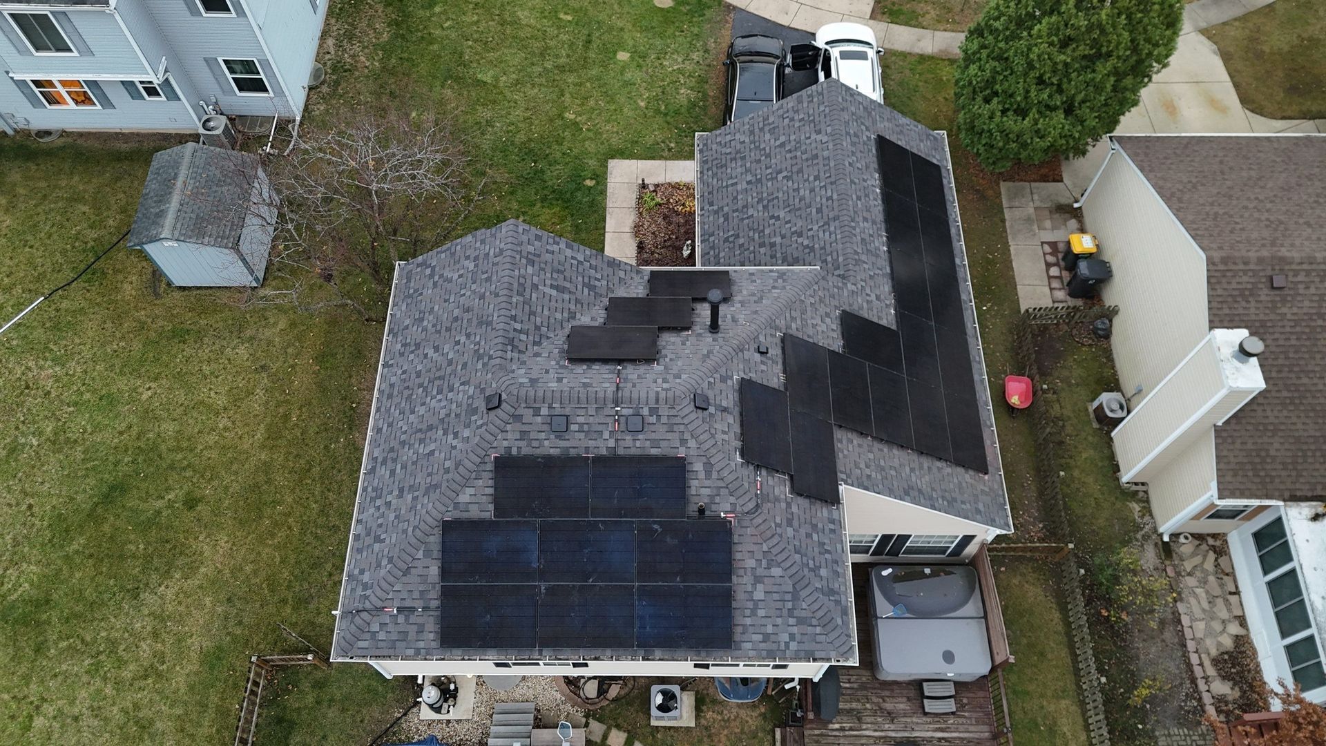 Aerial view of a house with solar panels on the roof, surrounded by grass, other houses and a shed.