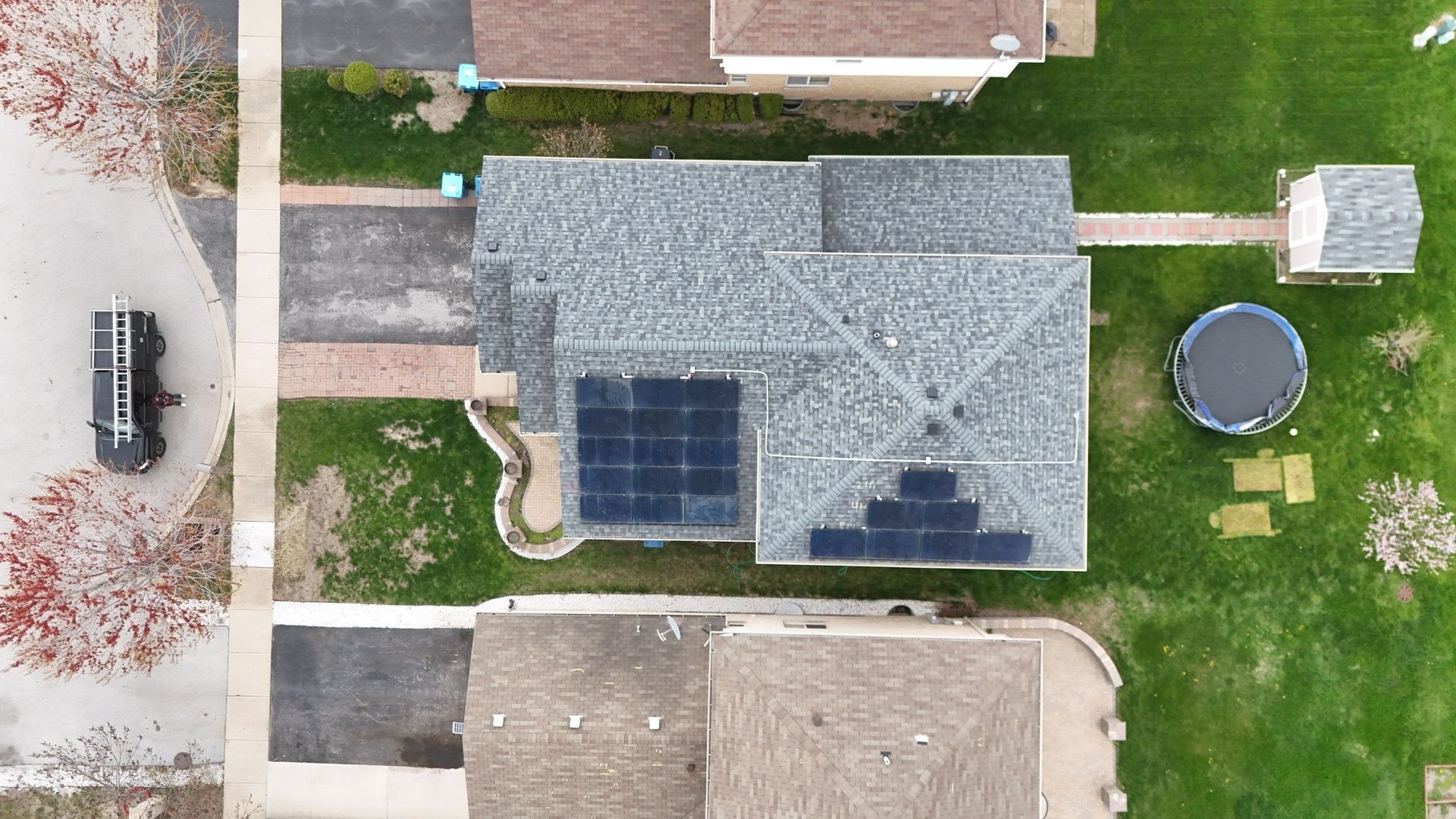 Overhead view of house with solar panels, a car in the driveway, a trampoline, and a shed in a grassy yard.