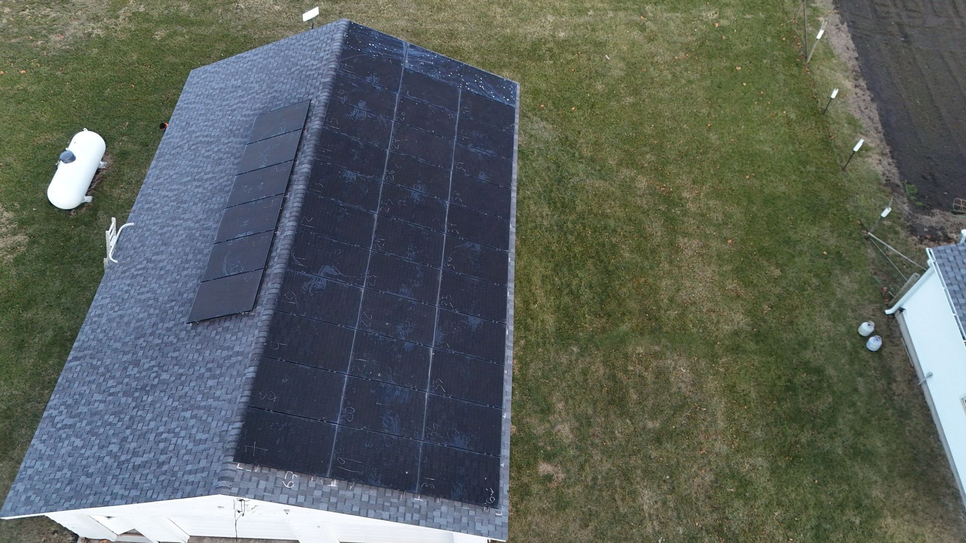 Solar panels on a gray shingled roof; propane tank and lawn in the background.