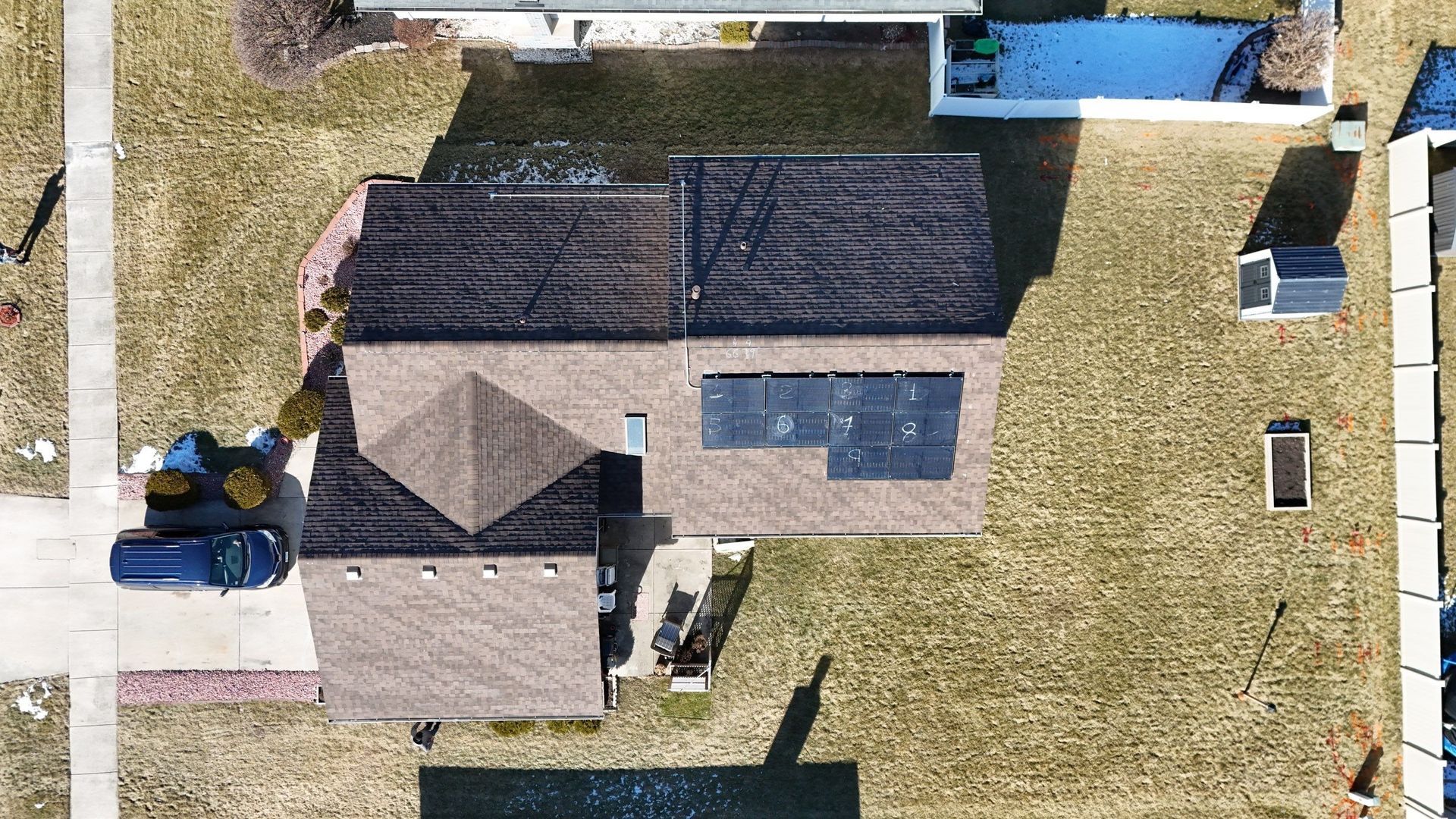 Overhead view of a house with solar panels on the roof, blue car in driveway, and a small shed in the yard.