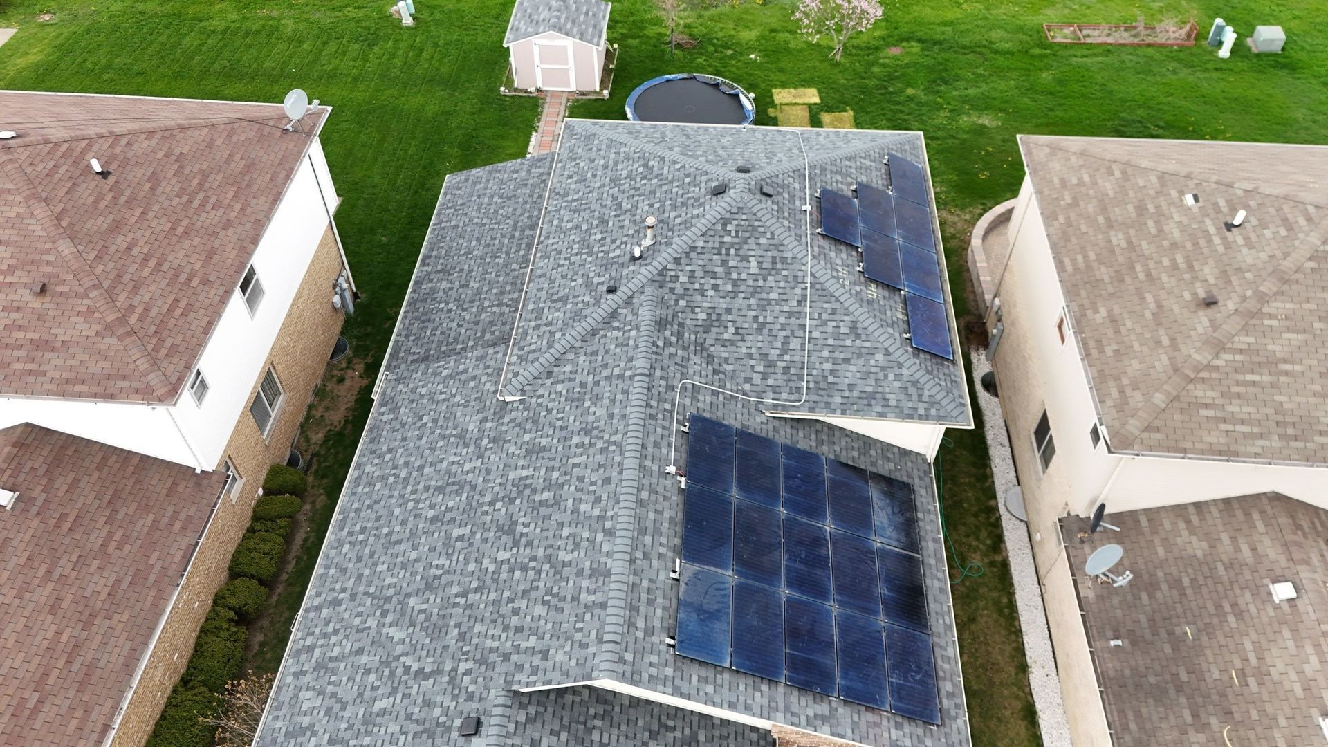 Overhead view of a house with solar panels on its roof, flanked by neighboring houses and a grassy backyard.