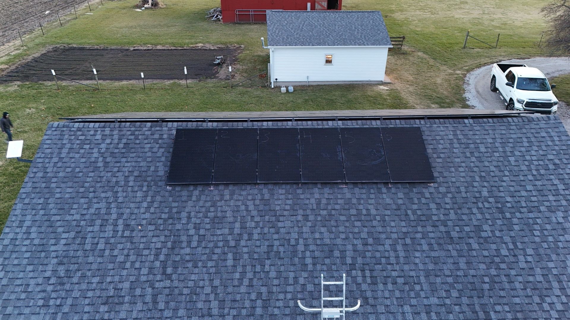 Solar panels on a dark roof of a house in a rural setting with a truck and outbuildings.