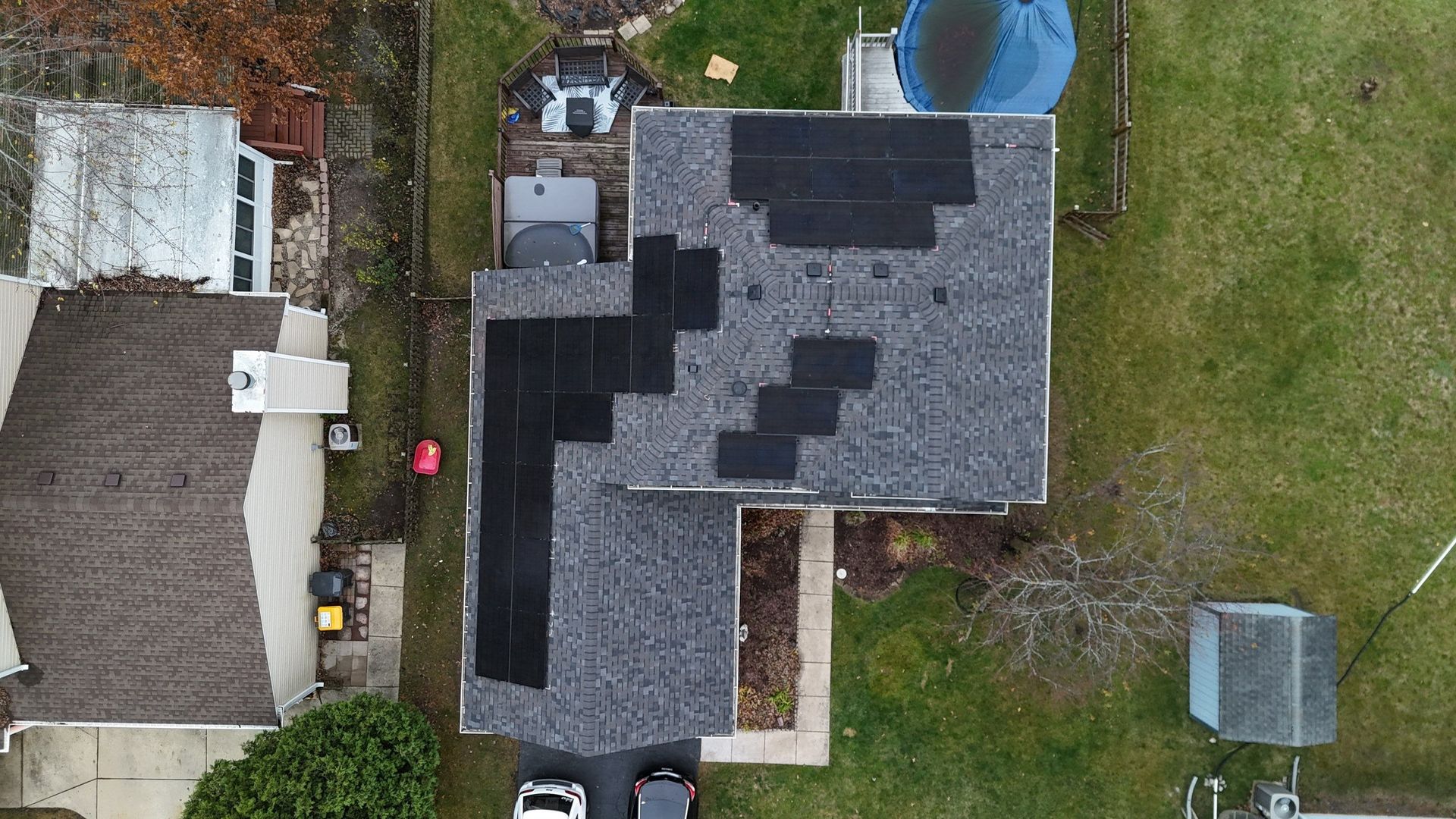 Overhead view of a house roof with installed solar panels. Gray roof, black panels, and surrounding yard.