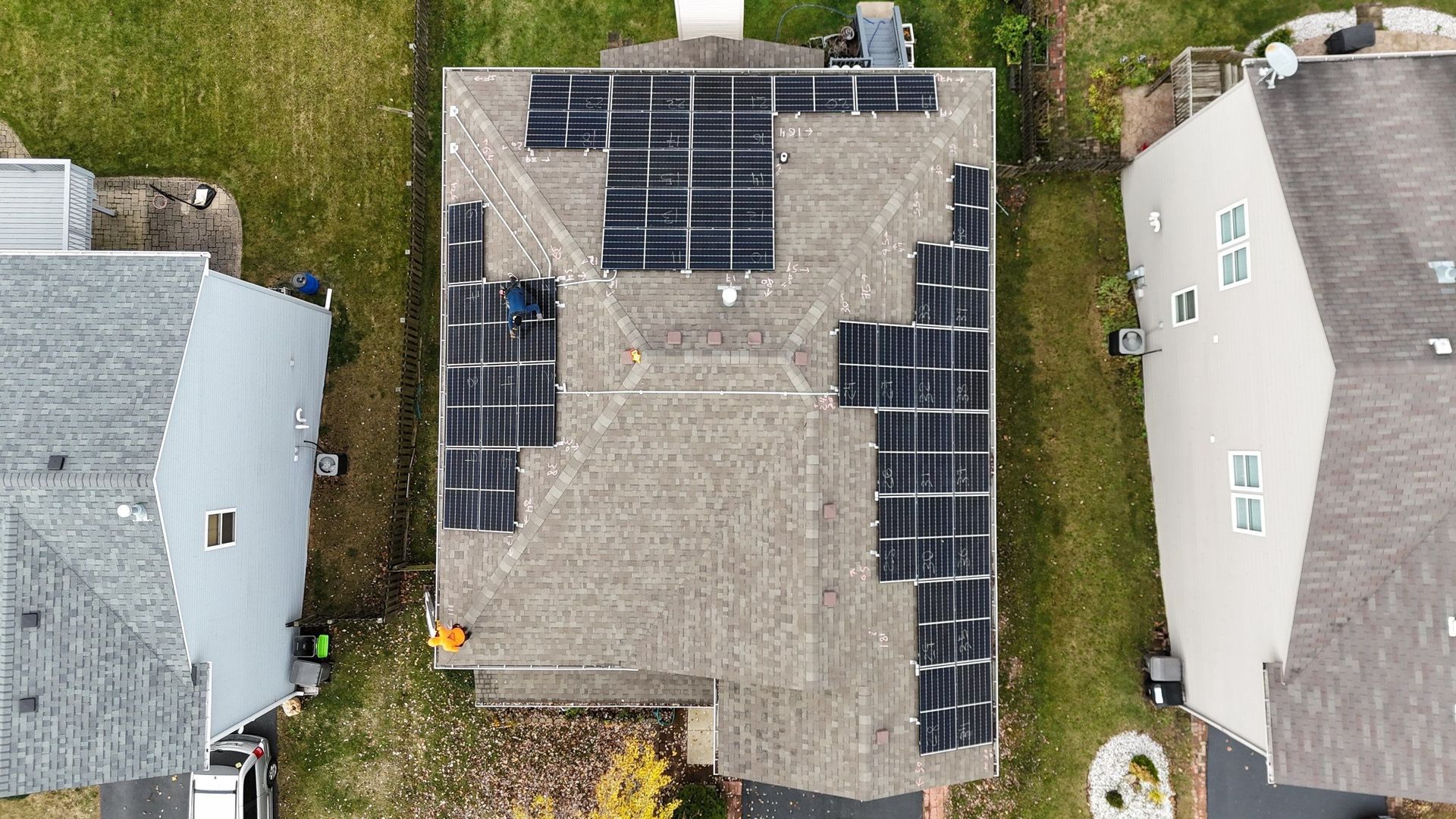 Aerial view of a house with solar panels being installed on its gray shingle roof. Houses surround it on both sides.