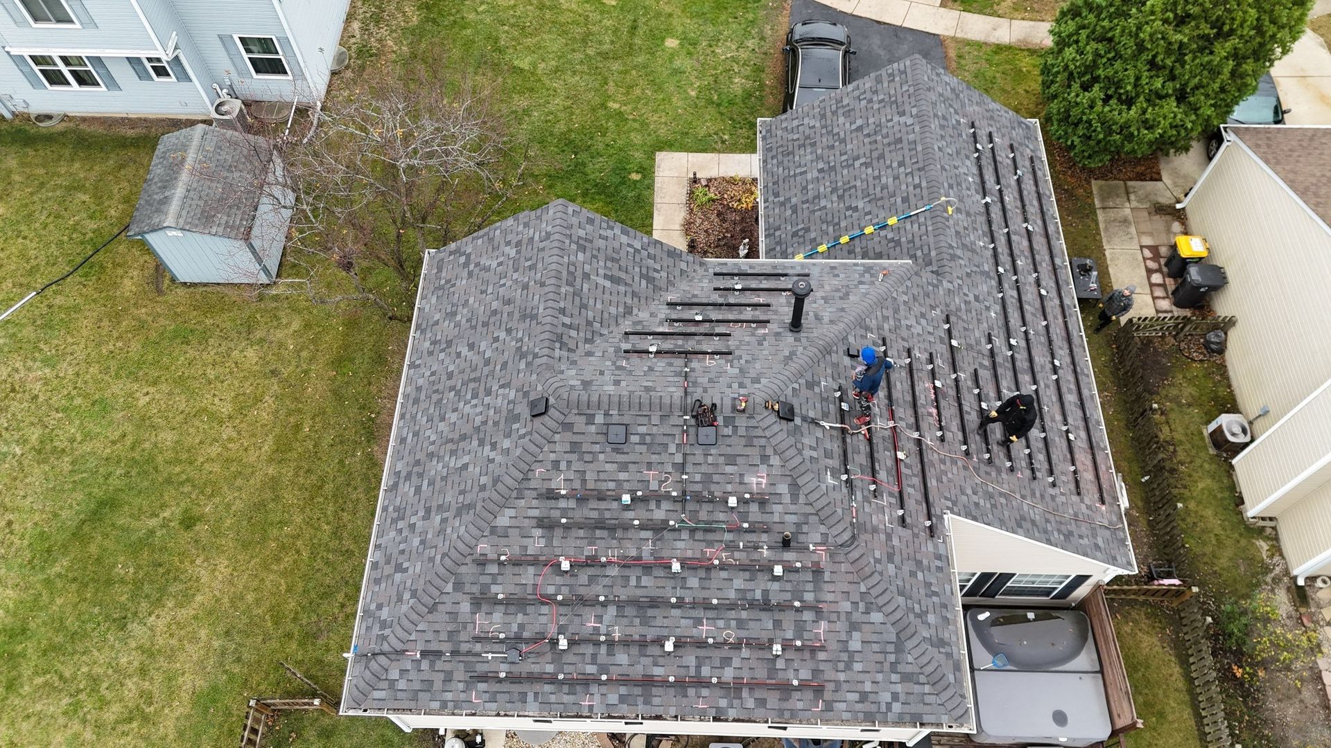 Aerial view of roofers installing solar panels on a gray shingle roof in a suburban setting.