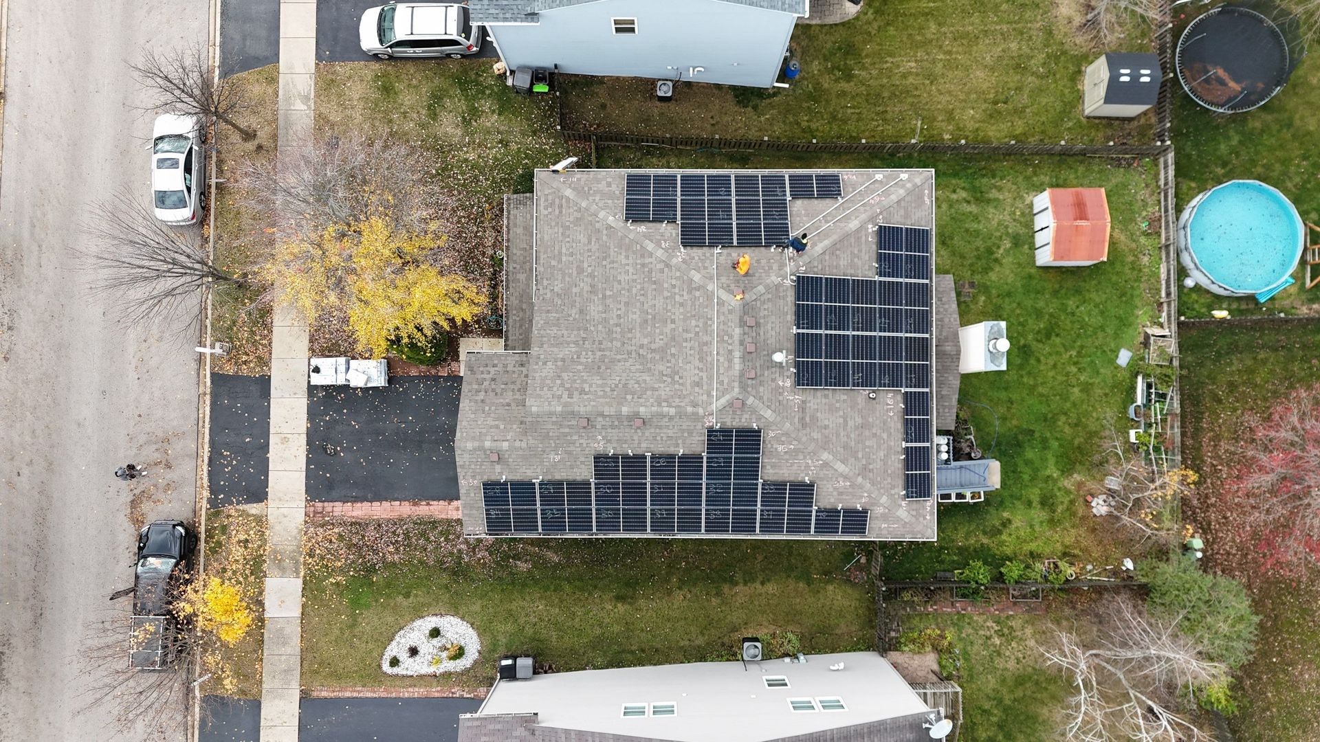 Overhead view of a house with solar panels installed on the roof, surrounded by grass, trees, and neighboring houses.