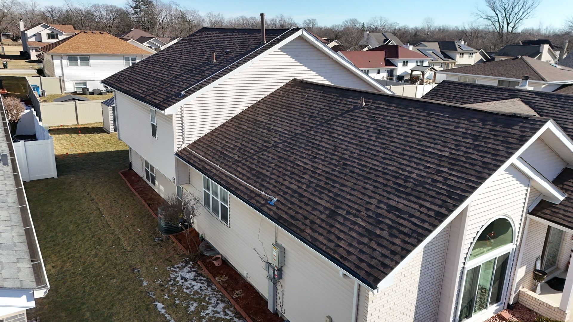 Residential house with dark brown shingled roof. Exterior walls are light tan. Winter setting, houses in background.