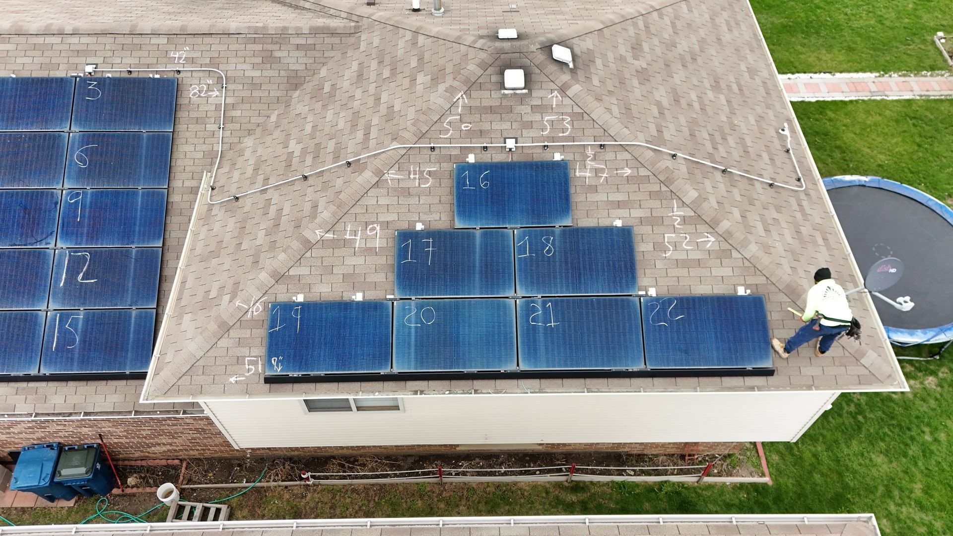 Aerial view of a roof with solar panels and a worker; green grass and trampoline in the yard.